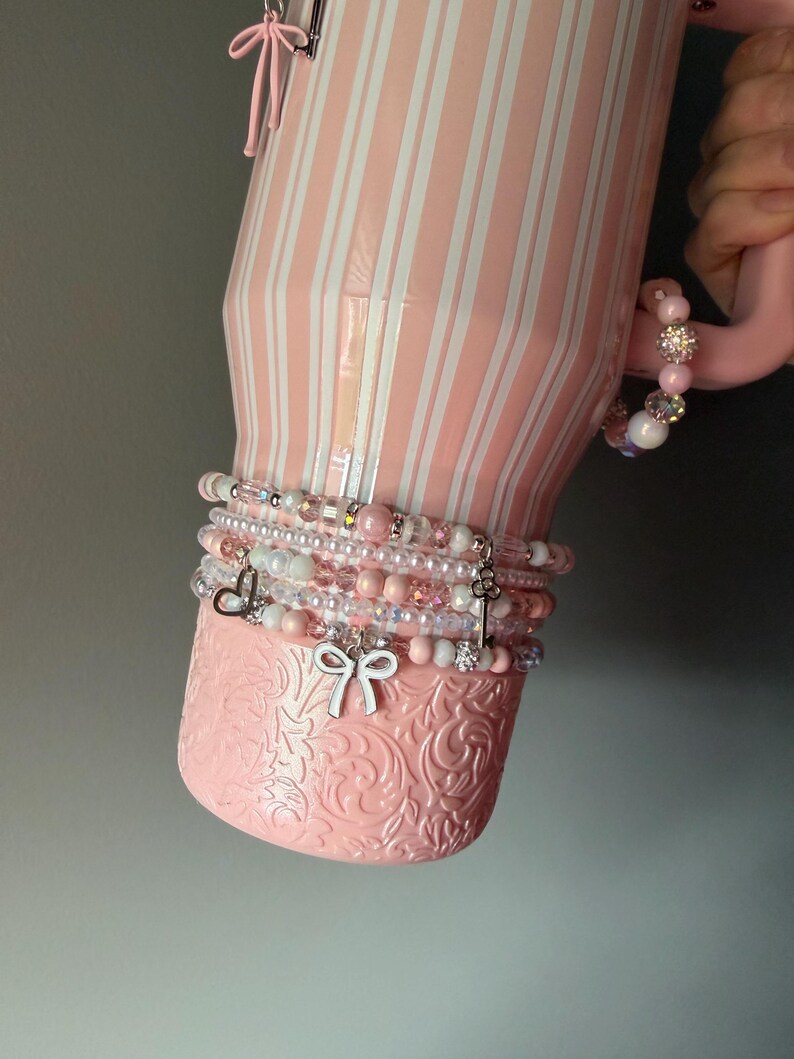 Pu&ograve; includere: Un bicchiere rosa chiaro con strisce verticali bianche e una base floreale testurizzata. Il bicchiere &egrave; decorato con una pila di braccialetti di perline con perle rosa e bianche, cristalli trasparenti e ciondoli argentati a forma di fiocco, cuore e chiave.
