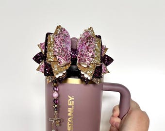 Op de afbeelding: Een mauve Stanley-beker met een decoratieve strik erop. De strik heeft lagen glinsterende gouden, paarse en roze stoffen. Een kralenbedel met een strik-hanger hangt aan de beker.