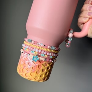 May include: A pink insulated tumbler with a decorative bicycle charm. The tumbler is adorned with several beaded bracelets in shades of pink, yellow, and blue. The bottom of the tumbler has a honeycomb pattern in yellow and orange.