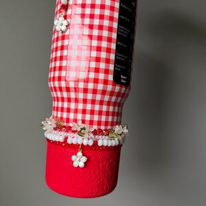 K&ouml;nnte beinhalten: Eine rot-wei&szlig; karierte Wasserflasche mit floralem Design. Die Flasche hat einen roten Boden mit gepr&auml;gtem Muster und ist mit floralen Perlenakzenten und einem Blumenanh&auml;nger verziert. Die Flasche ist 946 ml.