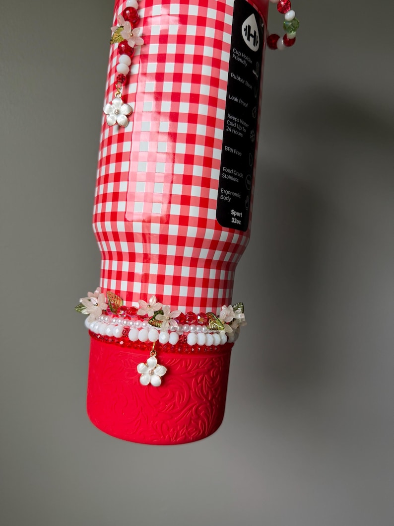 K&ouml;nnte beinhalten: Eine rot-wei&szlig; karierte Wasserflasche mit floralem Design. Die Flasche hat einen roten Boden mit gepr&auml;gtem Muster und ist mit floralen Perlenakzenten und einem Blumenanh&auml;nger verziert. Die Flasche ist 946 ml.