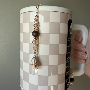 Può includere: Una tazza alta a scacchi con un ciondolo decorativo. Il ciondolo presenta una catena dorata con perline, una perlina di legno e un ciondolo a conchiglia. La tazza è bianca e beige con un motivo a scacchi. La tazza è tenuta da una persona.