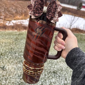 May include: A brown and gold stainless steel tumbler with a zebra print design and a brown handle. The tumbler has a brown glitter bow on top and gold beads around the bottom. The tumbler says "STANLEY" on the side.