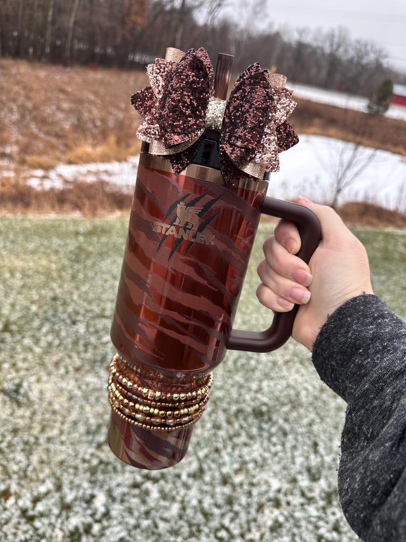 May include: A brown and gold stainless steel tumbler with a zebra print design and a brown handle. The tumbler has a brown glitter bow on top and gold beads around the bottom. The tumbler says "STANLEY" on the side.