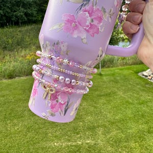 K&ouml;nnte beinhalten: Ein lilafarbener Becher mit Blumenmuster und einem goldenen Anh&auml;nger an der Henkel. Der Becher ist mit einer Reihe von rosa und goldenen Perlenarmb&auml;ndern verziert.