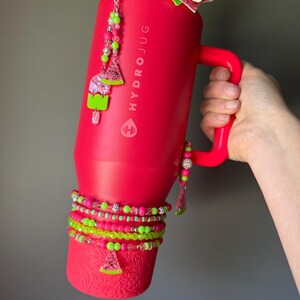 May include: A bright red HydroJug tumbler with a handle, featuring a large glittery bow on top. The tumbler is decorated with beaded charms shaped like watermelon slices and popsicles, and several beaded bracelets wrapped around the base.