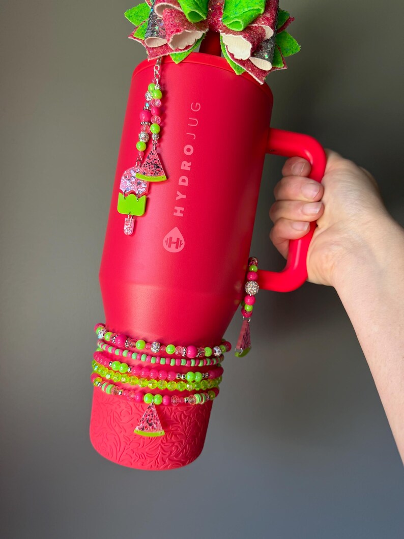 May include: A bright red HydroJug tumbler with a handle, featuring a large glittery bow on top. The tumbler is decorated with beaded charms shaped like watermelon slices and popsicles, and several beaded bracelets wrapped around the base.