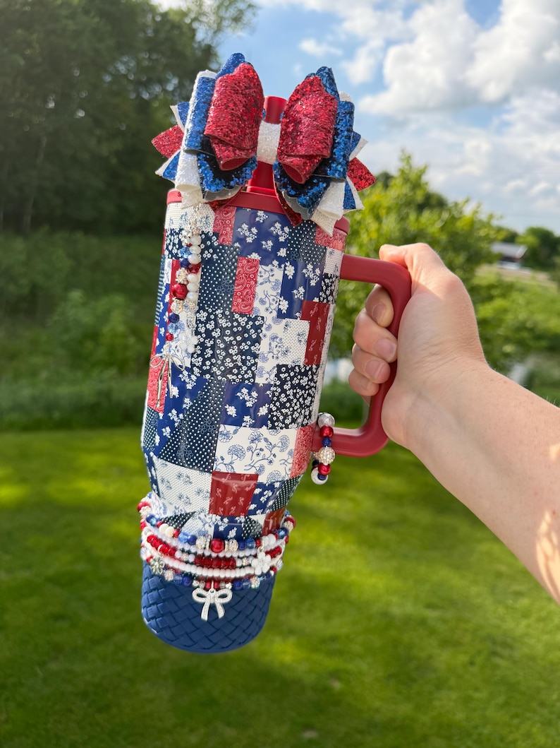 Puede incluir: Un vaso rojo, blanco y azul con un patr&oacute;n de patchwork de dise&ntilde;os florales y de lunares. El vaso tiene un asa roja, un adorno de lazo brillante en la parte superior y est&aacute; decorado con cuentas y un lazo blanco en la base del recipiente.