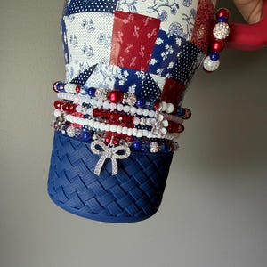 Puede incluir: Un vaso con un patr&oacute;n de patchwork en rojo, blanco y azul y una base de textura tejida azul. El vaso est&aacute; adornado con una pila de pulseras de cuentas en rojo, blanco y azul, con dijes que incluyen un lazo plateado y una flor blanca.