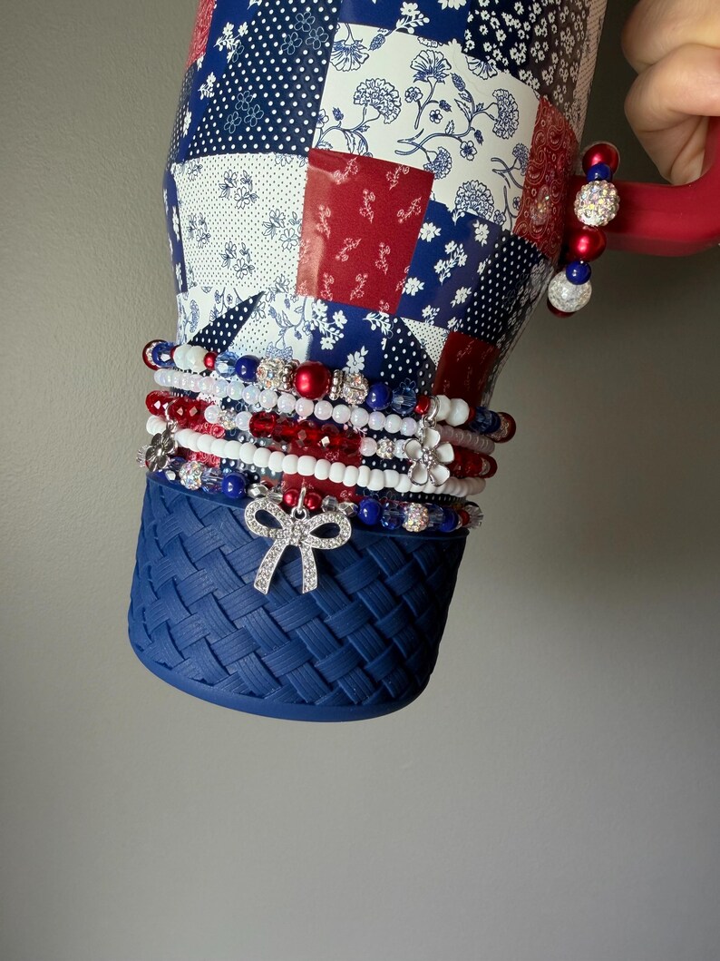 Puede incluir: Un vaso con un patr&oacute;n de patchwork en rojo, blanco y azul y una base de textura tejida azul. El vaso est&aacute; adornado con una pila de pulseras de cuentas en rojo, blanco y azul, con dijes que incluyen un lazo plateado y una flor blanca.