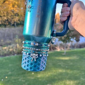 May include: A teal and blue insulated tumbler with a handle, adorned with beaded charms and bracelets. The tumbler has a decorative bow-shaped charm and a geometric patterned base. The background shows a grassy area and trees.