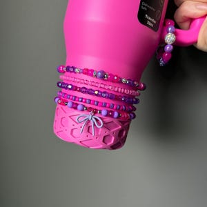 May include: A bright pink water bottle with a handle, adorned with several beaded bracelets in shades of pink and purple. The bracelets feature various bead sizes and textures, including some with sparkling accents. A small bow charm hangs from the bottom of the bottle.