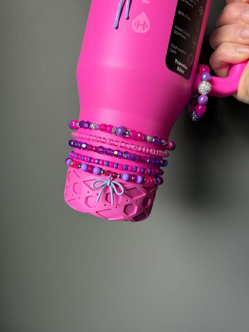 May include: A bright pink water bottle with a handle, adorned with several beaded bracelets in shades of pink and purple. The bracelets feature various bead sizes and textures, including some with sparkling accents. A small bow charm hangs from the bottom of the bottle.