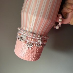 Pu&ograve; includere: Un bicchiere a righe rosa chiaro e bianco con una base testurizzata caratterizzata da un motivo floreale in rilievo. Il bicchiere &egrave; decorato con una serie di braccialetti di perline nei toni del rosa e del bianco, con ciondoli a forma di fiocco, cuore e chiave.