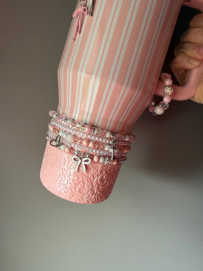 Pu&ograve; includere: Un bicchiere a righe rosa chiaro e bianco con una base testurizzata caratterizzata da un motivo floreale in rilievo. Il bicchiere &egrave; decorato con una serie di braccialetti di perline nei toni del rosa e del bianco, con ciondoli a forma di fiocco, cuore e chiave.