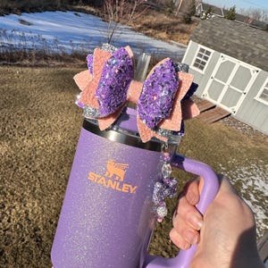 May include: A purple glitter tumbler with a silver lid and a purple handle. The tumbler has a "STANLEY" logo on the front. A pink and purple glitter bow with a silver ribbon is attached to the lid.