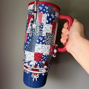 Puede incluir: Un vaso t&eacute;rmico alto con asa y tapa rojas, que presenta un patr&oacute;n de patchwork en rojo, blanco y azul. El vaso est&aacute; adornado con m&uacute;ltiples dijes de cuentas, incluyendo cuentas blancas, rojas y azules y un dije de lazo plateado en la base.