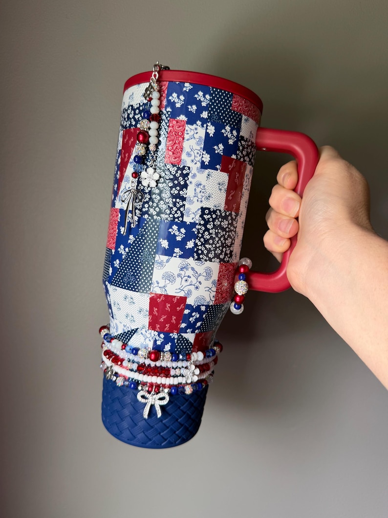 Puede incluir: Un vaso t&eacute;rmico alto con asa y tapa rojas, que presenta un patr&oacute;n de patchwork en rojo, blanco y azul. El vaso est&aacute; adornado con m&uacute;ltiples dijes de cuentas, incluyendo cuentas blancas, rojas y azules y un dije de lazo plateado en la base.