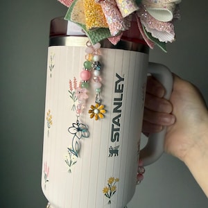 May include: A set of three ceramic mugs with a speckled, cream-colored glaze. Each mug features a different hand-painted floral design in shades of blue, green, and yellow. The mugs have a simple, rounded shape and a comfortable handle.