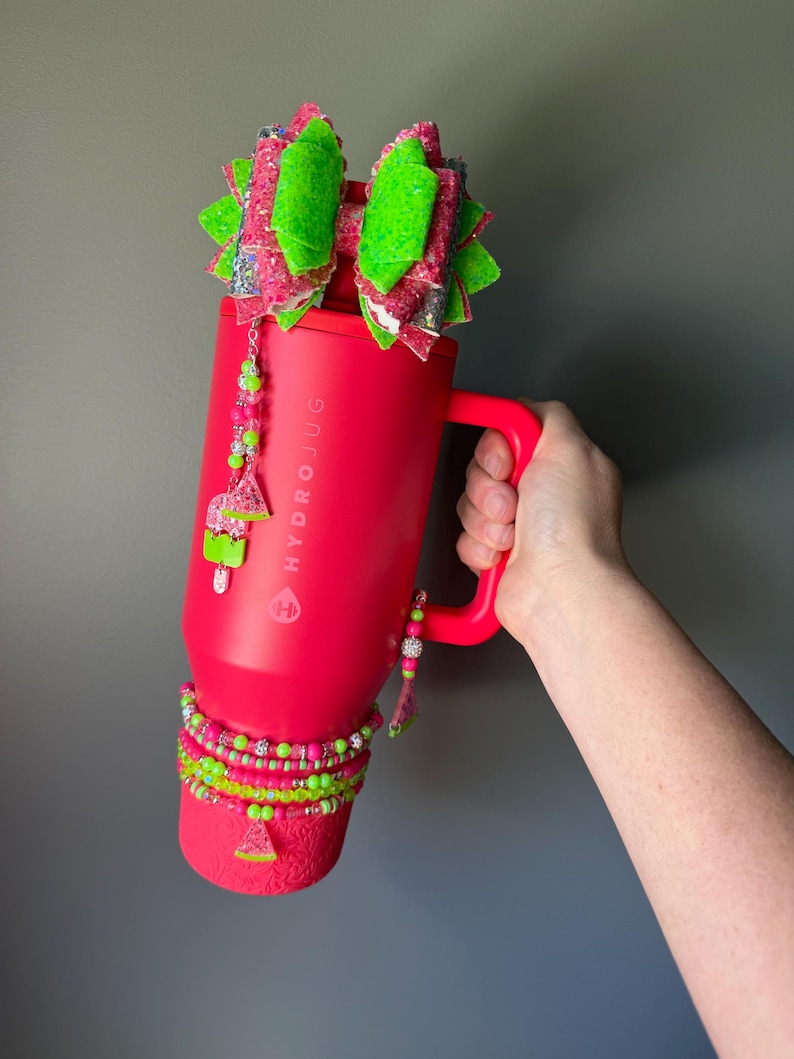 May include: A bright red HydroJug tumbler featuring a large, multi-layered glittery bow in neon green and pink attached to the lid. The tumbler is decorated with beaded charms, including watermelon slices, and a stack of matching neon beaded bracelets.