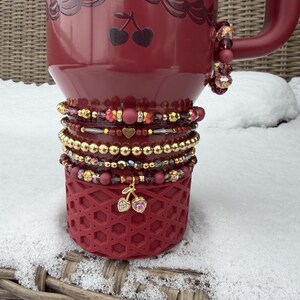 May include: A burgundy travel mug with a decorative cherry design and a stack of beaded bracelets. The bracelets feature gold, red, and pink beads, with a cherry charm. The mug is sitting on a woven surface.