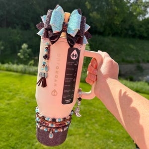 Puede incluir: Una botella de agua HydroJug rosa claro con asa, decorada con un gran lazo con detalles de purpurina turquesa y negra. La botella est&aacute; adornada con pulseras de cuentas y abalorios, incluyendo turquesa y una bota plateada.