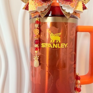 May include: A red Stanley mug with an orange handle, adorned with a decorative bow and beaded charms. The bow features glittery gold, yellow, and red accents. The mug has the Stanley logo in gold and a floral pattern.