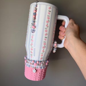 May include: A large white mug with a floral and striped pattern, adorned with beaded charms and a pink knitted cozy. The mug has a white handle and is decorated with pink and blue beads and charms.