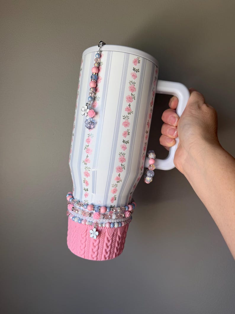 May include: A large white mug with a floral and striped pattern, adorned with beaded charms and a pink knitted cozy. The mug has a white handle and is decorated with pink and blue beads and charms.
