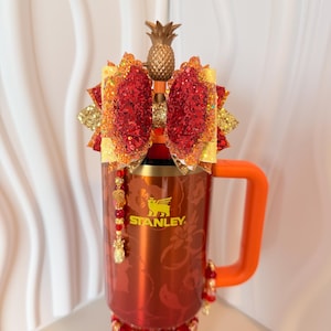 May include: A red Stanley mug with an orange handle, adorned with a gold pineapple topper and a red bow with gold glitter accents. The mug is decorated with floral patterns and a beaded embellishment.
