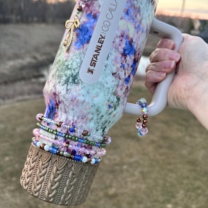 May include: A white and blue floral patterned stainless steel travel mug with a white handle. The mug has a gold braided band around the bottom and a gold charm with a bow hanging from the top. The mug is labeled "STANLEY GO CALLA".
