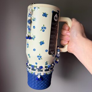 Op de afbeelding: Een 946 ml cr&egrave;me-kleurige ge&iuml;soleerde beker met een blauw bosbessenpatroon, een donkerblauwe basis met gevlochten textuur en decoratieve kralenbedels. De beker heeft een zwart label met witte tekst die kenmerken zoals bekerhouder-vriendelijk, lekvrij en vaatwasserbestendig beschrijft.