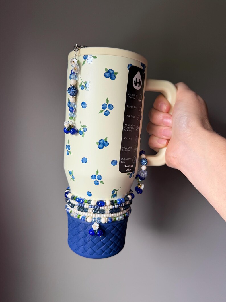 Op de afbeelding: Een 946 ml cr&egrave;me-kleurige ge&iuml;soleerde beker met een blauw bosbessenpatroon, een donkerblauwe basis met gevlochten textuur en decoratieve kralenbedels. De beker heeft een zwart label met witte tekst die kenmerken zoals bekerhouder-vriendelijk, lekvrij en vaatwasserbestendig beschrijft.