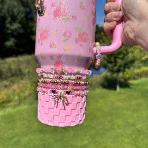 May include: Pink Stanley tumbler with floral design and the text "STANLEY LOVESHACK FANCY". The tumbler is accessorized with multiple beaded bracelets and a pink basket-weave bottom. The handle is pink.