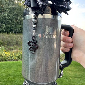 Peut inclure: Un gobelet Stanley gris avec une poignée noire, orné d'un nœud noir, d'accents perlés et d'un charme de serpent. Le gobelet est orné de plusieurs couches de perles noires et argentées autour de la base.