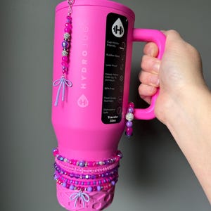 May include: A pink HydroJug Traveler 32oz water bottle with a handle, adorned with beaded bracelets and charms in shades of pink and purple. The bottle features the HydroJug logo and product information.