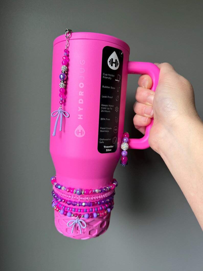 May include: A pink HydroJug Traveler 32oz water bottle with a handle, adorned with beaded bracelets and charms in shades of pink and purple. The bottle features the HydroJug logo and product information.