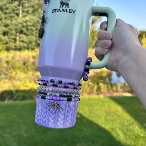 May include: A Stanley mug with a light green to purple gradient, decorated with black, purple, and clear beaded accents. The base has a knitted purple sleeve and a bow. The text "Witches Brew" is spelled out in beads.