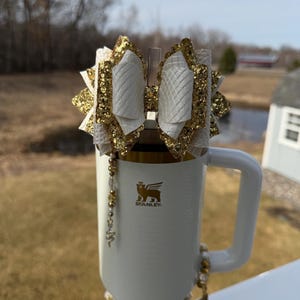 Könnte beinhalten: Ein weißer Stanley-Becher mit goldenem Rand und Griff, verziert mit einer gold-weißen Schleife. Die Schleife hat einen glitzernden Goldrand und eine gesteppte weiße Mitte. Ein Perlenanhänger hängt am Becher, und ein Perlenarmband umschließt den Boden.
