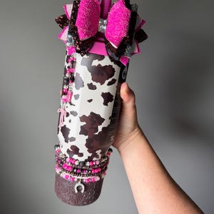 May include: A cylindrical tumbler featuring a white and brown cow print pattern. The top is adorned with a large, multi-layered hot pink and dark brown glitter bow. A beaded charm hangs from the side, and a beaded bracelet with a horseshoe charm.