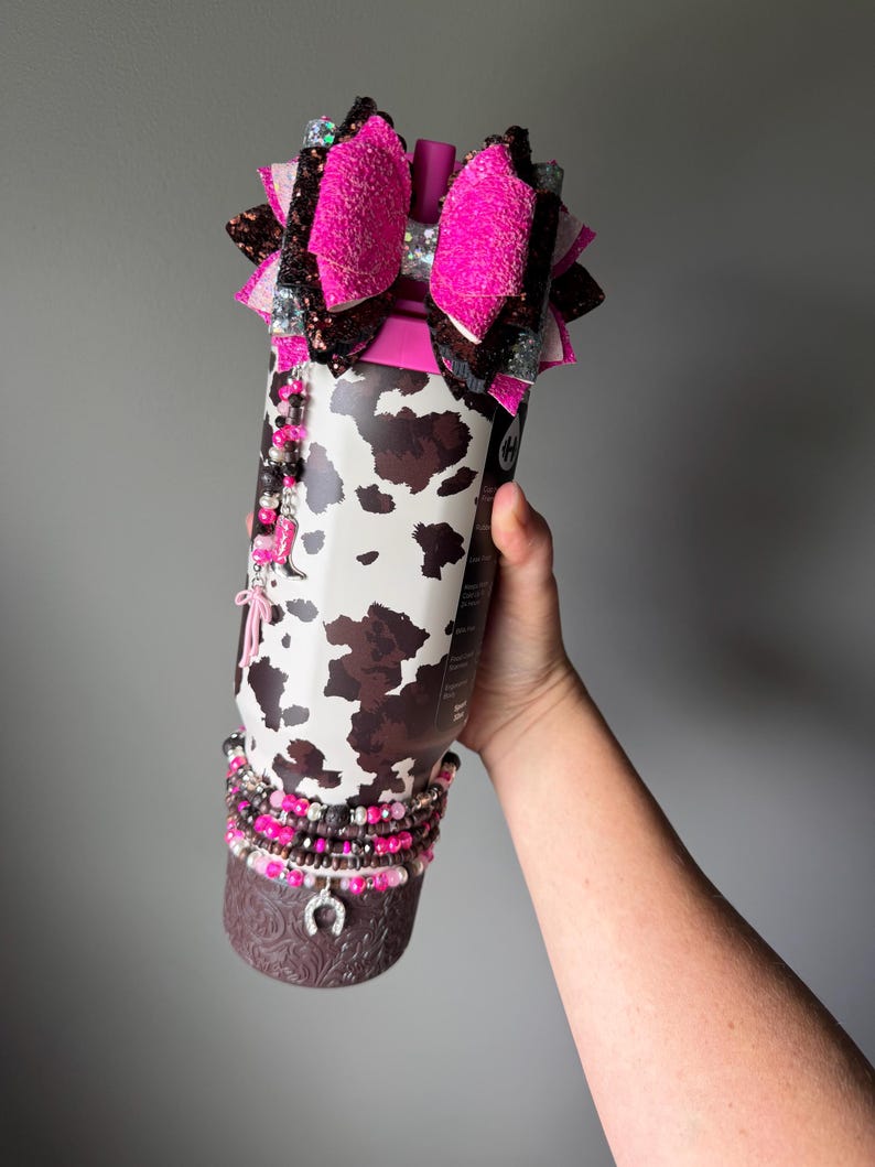 May include: A cylindrical tumbler featuring a white and brown cow print pattern. The top is adorned with a large, multi-layered hot pink and dark brown glitter bow. A beaded charm hangs from the side, and a beaded bracelet with a horseshoe charm.