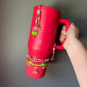 May include: A bright pink HydroJug tumbler with a handle, decorated with a beaded charm featuring watermelon and popsicle shapes. The base is wrapped in multiple layers of neon pink and lime green beaded bracelets with a small watermelon charm.