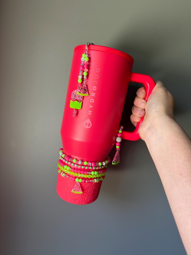 May include: A bright pink HydroJug tumbler with a handle, decorated with a beaded charm featuring watermelon and popsicle shapes. The base is wrapped in multiple layers of neon pink and lime green beaded bracelets with a small watermelon charm.