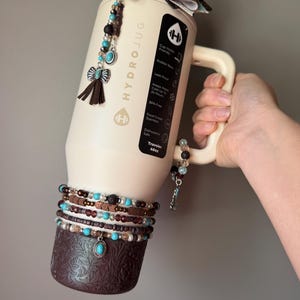 May include: A cream-colored HydroJug water bottle with a decorative bow and beaded accents. The bottle features a handle and is adorned with multiple beaded bracelets and a decorative bottom. The bottle is labeled "HYDROJUG" and has a capacity of 40oz.