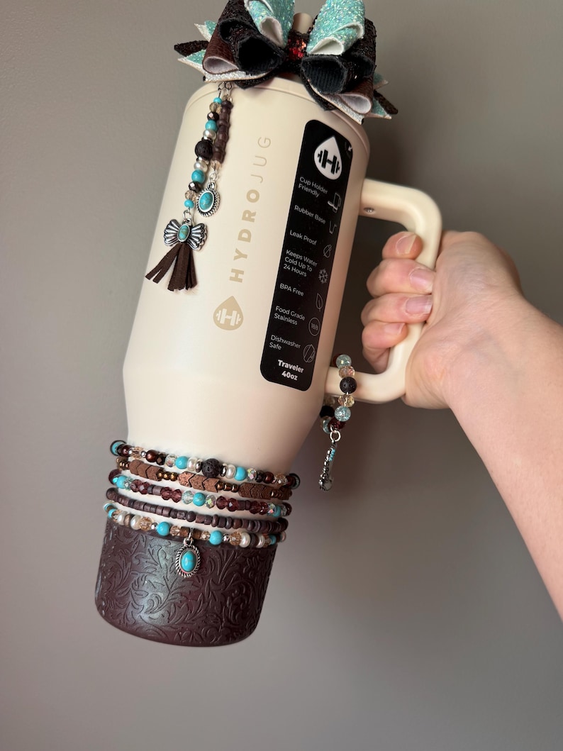 May include: A cream-colored HydroJug water bottle with a decorative bow and beaded accents. The bottle features a handle and is adorned with multiple beaded bracelets and a decorative bottom. The bottle is labeled "HYDROJUG" and has a capacity of 40oz.