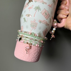 Puede incluir: Un vaso blanco con asa rosa y un estampado de vacaciones tropicales con flamencos rosas, palmeras y coches antiguos. El vaso est&aacute; decorado con varias pulseras de cuentas en tonos rosa y verde, con peque&ntilde;os dijes met&aacute;licos de flamencos y hojas.