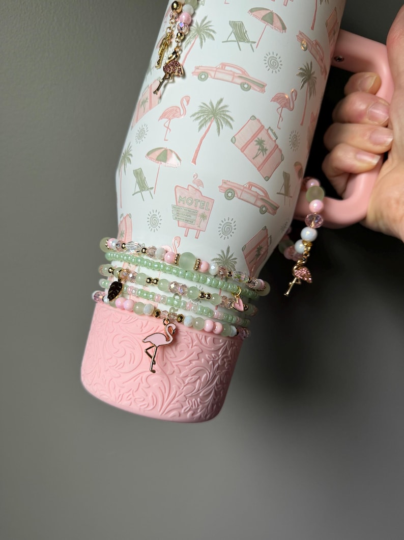 Puede incluir: Un vaso blanco con asa rosa y un estampado de vacaciones tropicales con flamencos rosas, palmeras y coches antiguos. El vaso est&aacute; decorado con varias pulseras de cuentas en tonos rosa y verde, con peque&ntilde;os dijes met&aacute;licos de flamencos y hojas.