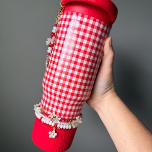 K&ouml;nnte beinhalten: Ein rot-wei&szlig; karierter Becher mit rotem Deckel und Boden. Der Becher ist mit einem floralen Perlenanh&auml;nger und einem wei&szlig;-roten Perlenarmband verziert. Der Becher wird von einer Person gehalten.