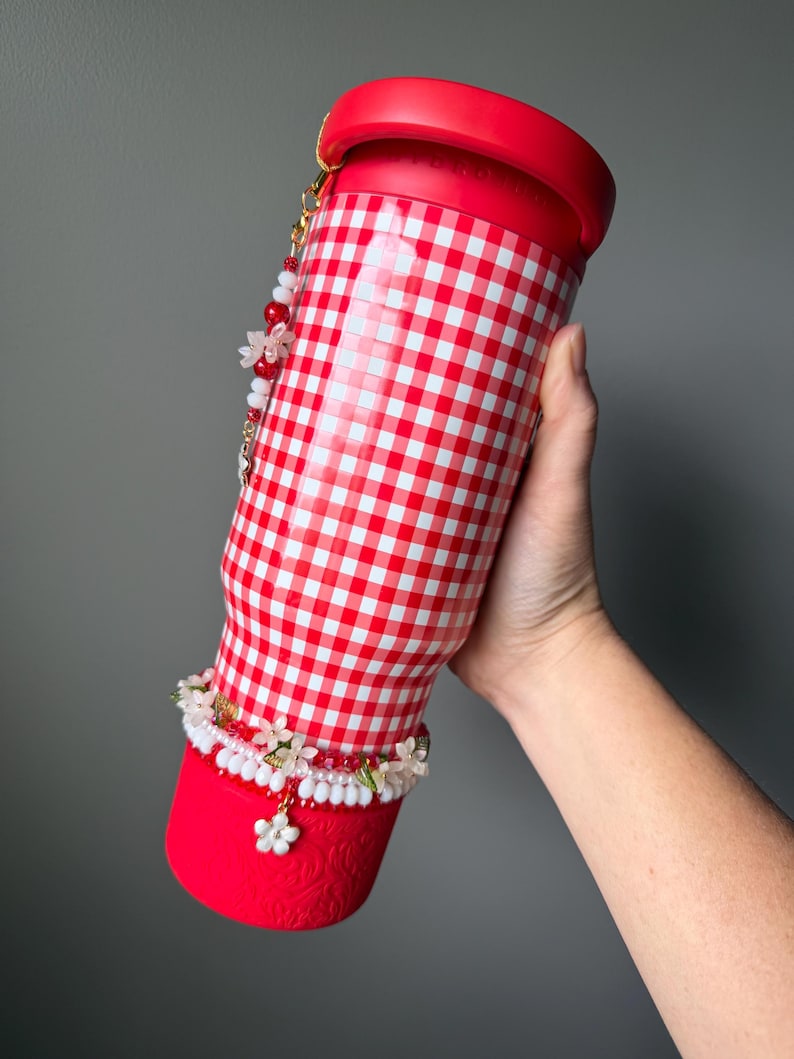 K&ouml;nnte beinhalten: Ein rot-wei&szlig; karierter Becher mit rotem Deckel und Boden. Der Becher ist mit einem floralen Perlenanh&auml;nger und einem wei&szlig;-roten Perlenarmband verziert. Der Becher wird von einer Person gehalten.