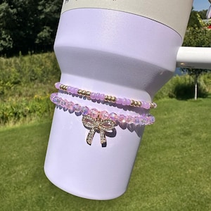 K&ouml;nnte beinhalten: Ein lilafarbener 40 oz Tumbler in limitierter Farbe. Zwei rosafarbene und goldene Perlenarmb&auml;nder mit einem Strass-Schleifenanh&auml;nger sind um den Tumbler gewickelt.